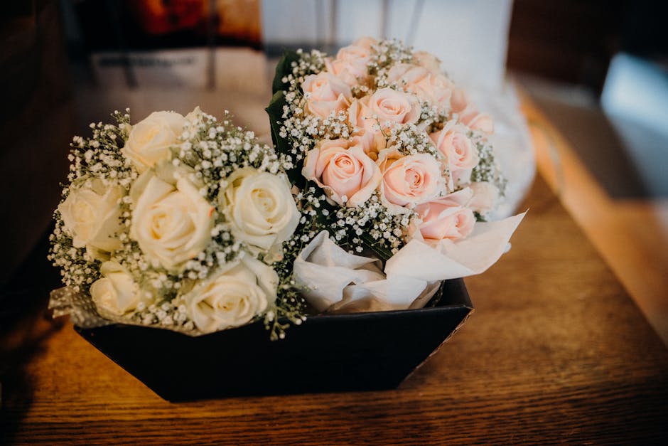 A black rectangular box containing two floral bouquets placed on a wooden surface. One bouquet features white roses surrounded by small white baby's breath flowers and greenery, arranged in a compact,