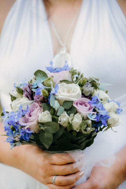 A bride holding a floral bouquet arrangement composed of white roses, light purple roses, and small blue flowers, accented with green foliage. The bouquet is wrapped with white tulle and held in both