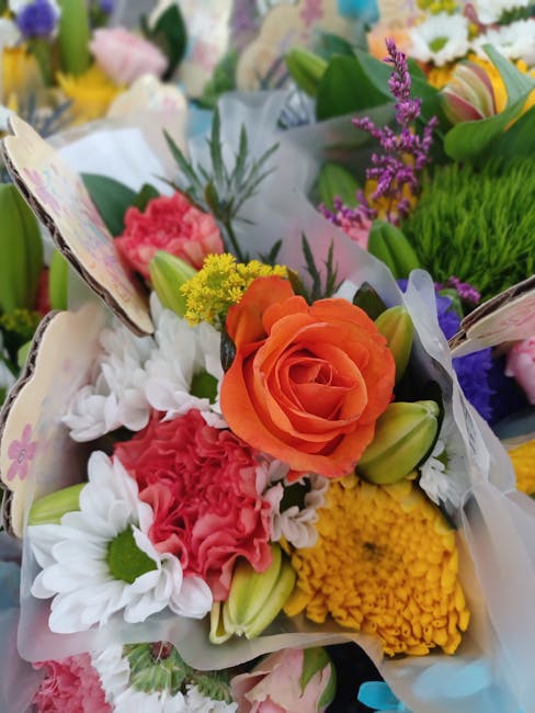 A vibrant floral arrangement featuring an orange rose as the focal point, surrounded by white daisies with green centers, pink carnations, yellow chrysanthemums, and purple statice, all wrapped in whi