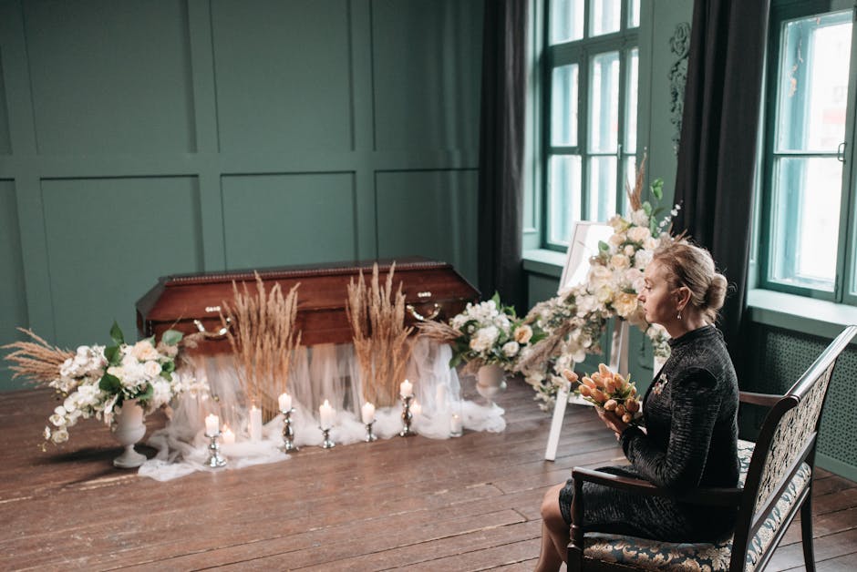 A woman seated in a floral-scented room holding a bouquet of fresh flowers, with a arrangement of white and blush flowers and green foliage in front of her. The arrangement is displayed on a tripod st