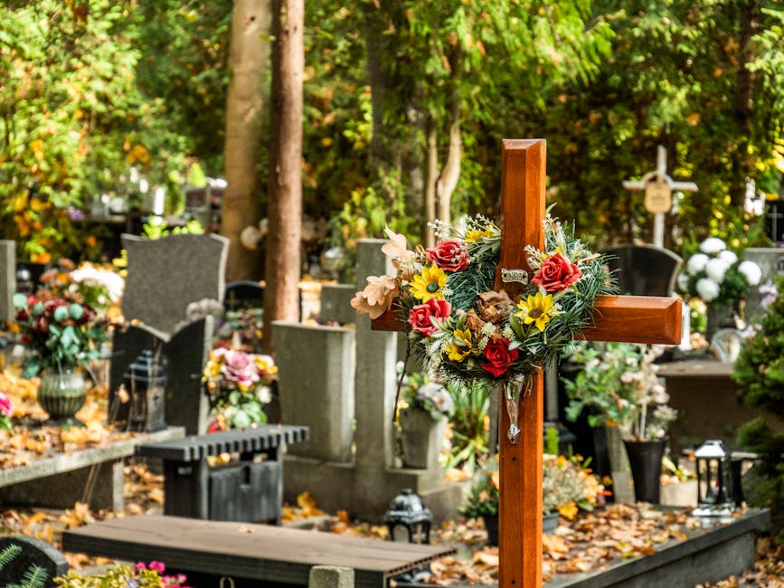A wooden cross decorated with a floral arrangement featuring red roses, yellow daisies, pale pink flowers, and green foliage, situated in a cemetery surrounded by various gravestones, some with potted